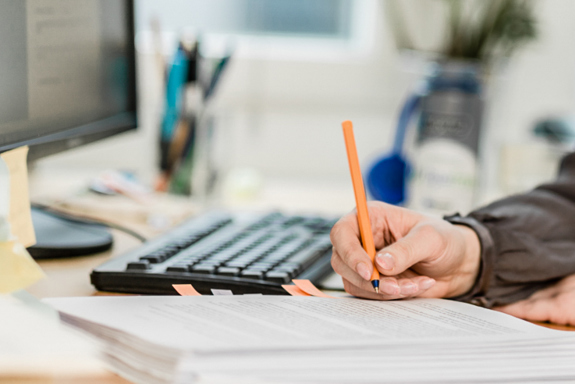 Fotoet viser en person som sitter ved en PC og har en bunke med papirer foran seg.