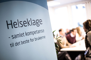 En roll up med teksten "Helseklage – samlet kompetanse til det beste for brukerne". I bakgrunnen skimtes flere personer. Illustrasjonsfoto: Ådne Sinnes.