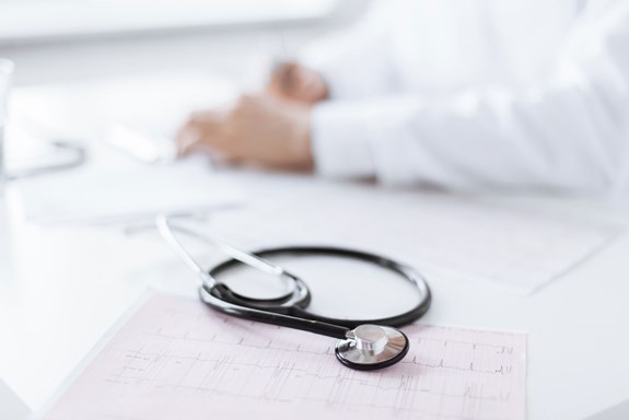 Et stetoskop ligger oppå noen papirer på et bord, bak bordet sitter en lege. Foto: Mostphotos.