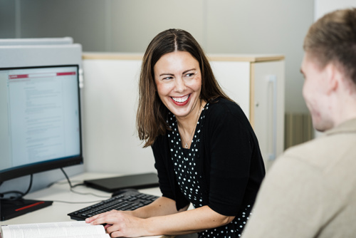Bildet viser en kvinne som sitter ved en kontorpult og smiler mot en kollega.