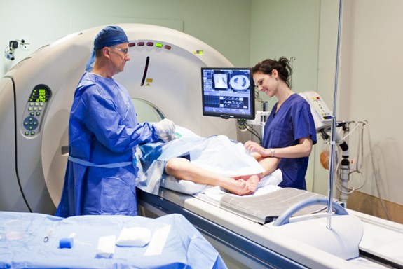 Bildet syner ein pasient som ligg på ei MR-maskin. To personar som er helsepersonell står ved sidan av.