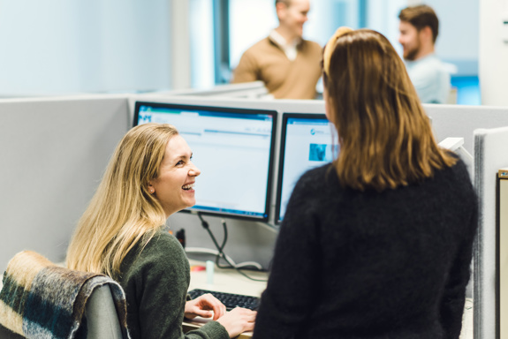 To personer snakker sammen i et kontorlandskap. Flere personer er i bakgrunnen. Foto: Cecilie Bannow.