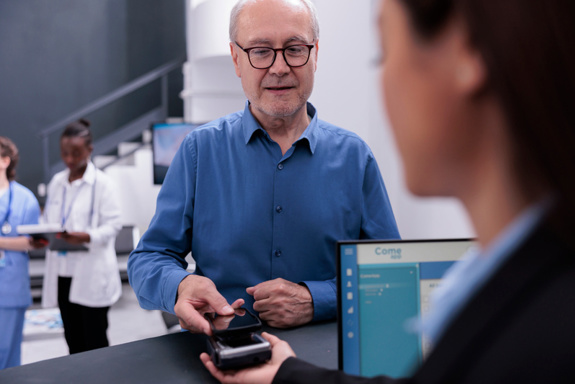 Bildet viser en pasient som betaler med telefonen sin på kortautomaten i en resepsjon. En person holder i kortautomaten. I bakgrunnen står to helsepersonell. Foto: Mostphotos.